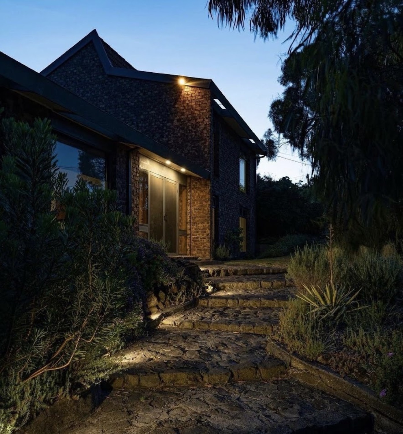 Stone steps and garden beds lit beside a brick home at dusk